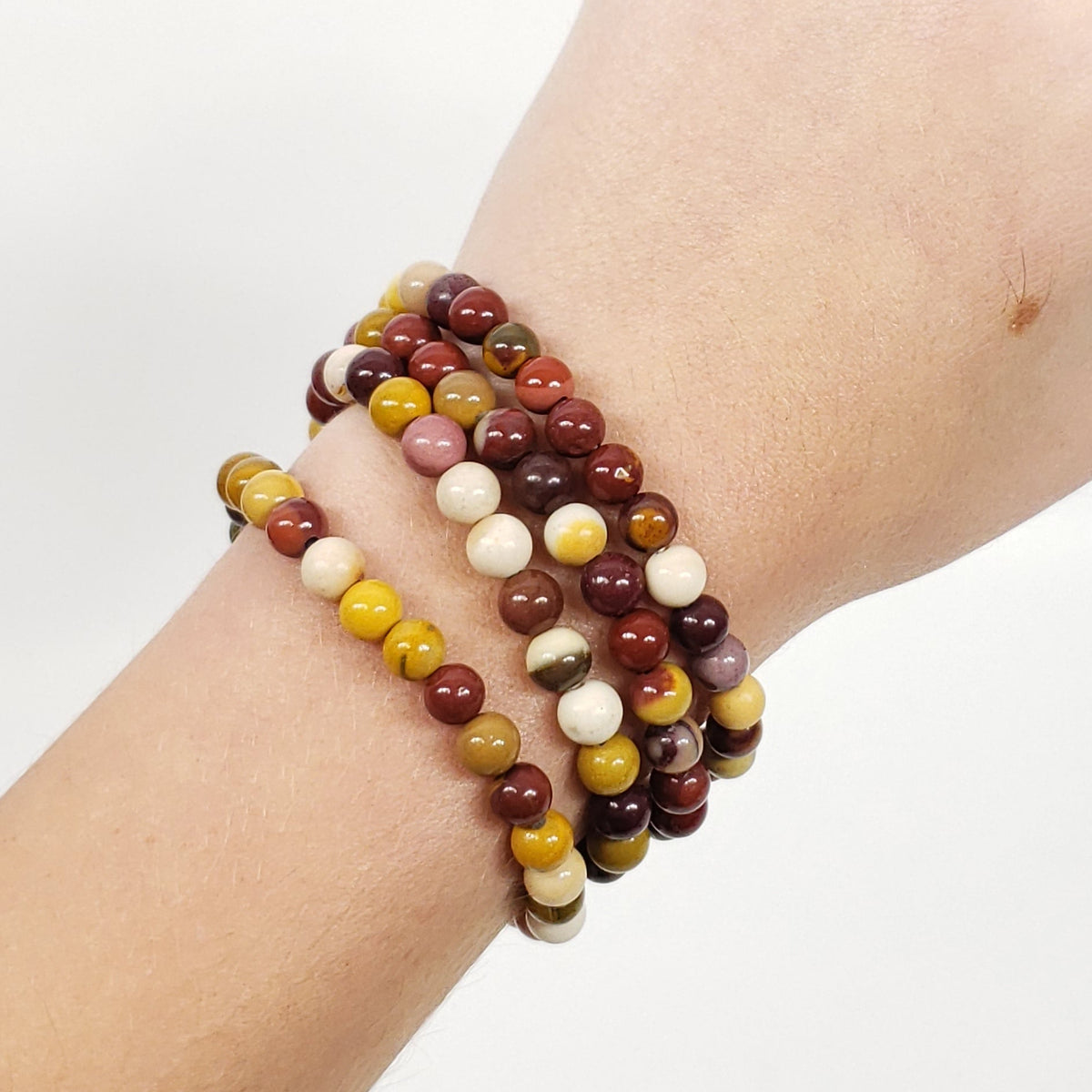 A close-up of a person's wrist wearing three 4mm beaded mookaite jasper stretch bracelets in earthy brown, yellow, cream, and burgundy tones, with a gold ring on the finger, against a plain white background.