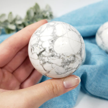 A hand holds a polished white stone sphere with gray and black veining, with a blue fabric and blurred greenery in the background.
