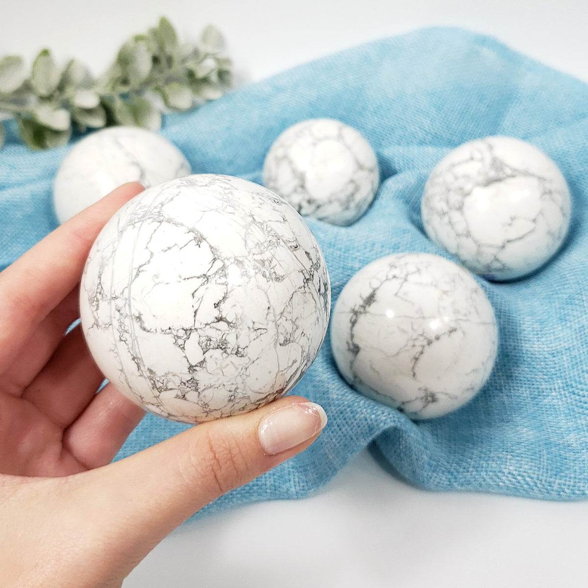 A hand holds a white marble-patterned decorative sphere in the foreground, with four matching spheres arranged on a light blue fabric backdrop. A small green leafy sprig appears in the top left corner.