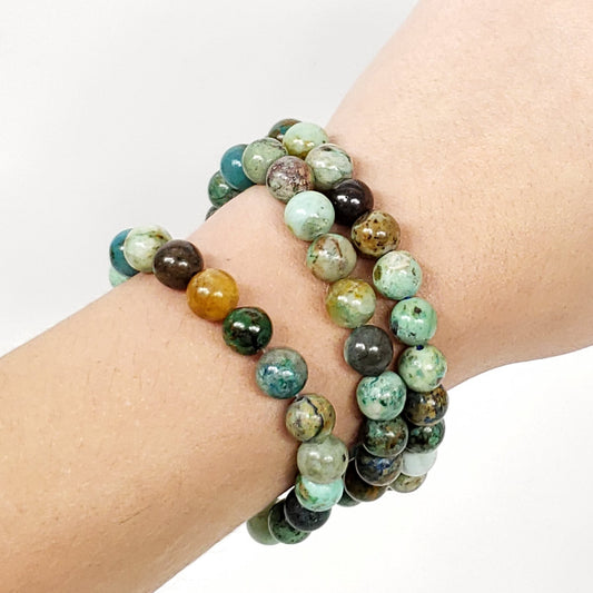 A close-up of a person's wrist wearing three beaded Chrysocolla bracelets made of polished round stones in green, brown, black, and amber tones, against a plain white background.
