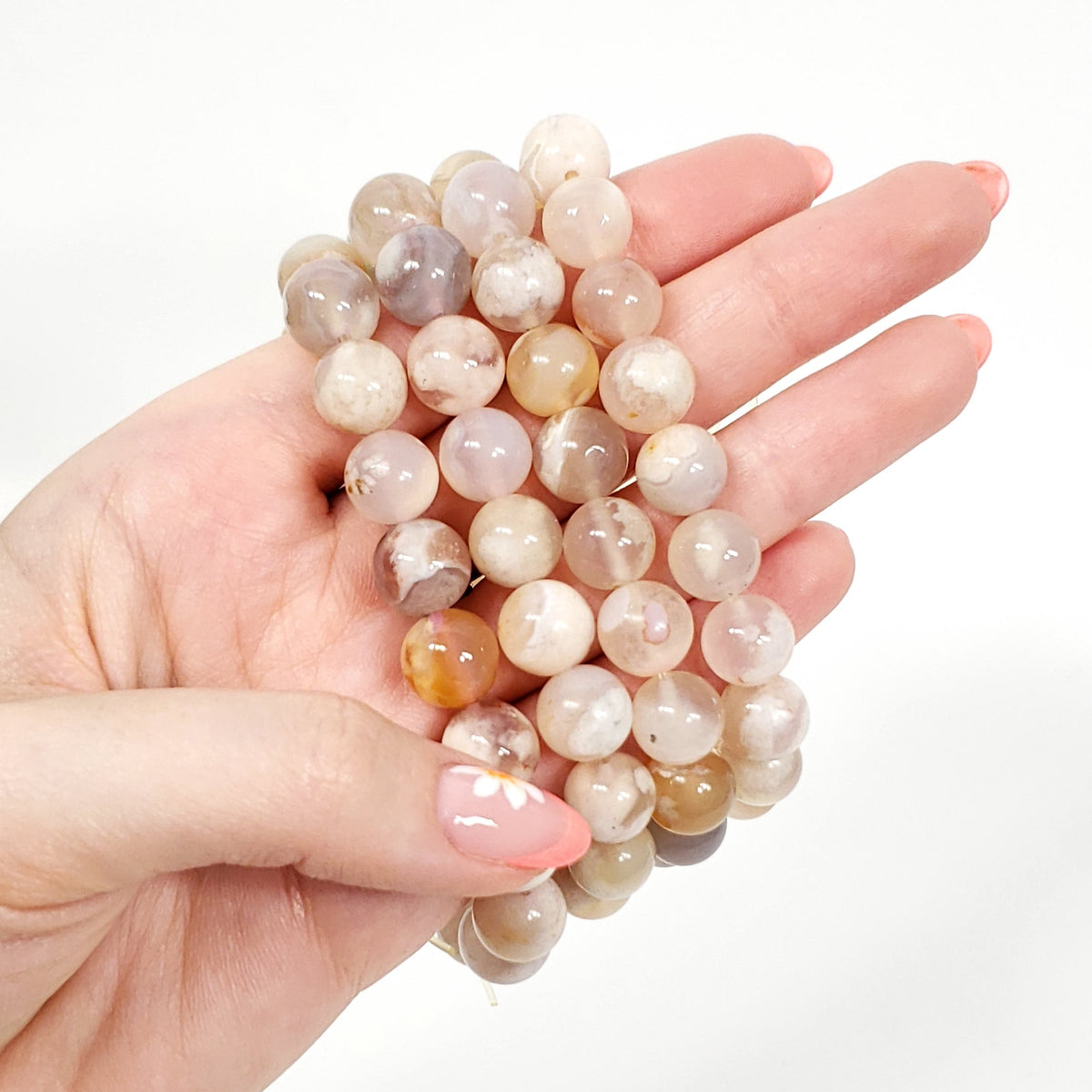 A hand holding a flower agate beaded bracelet made of smooth, polished round beads in soft neutral tones of beige, cream, light brown, and peach against a clean white background.