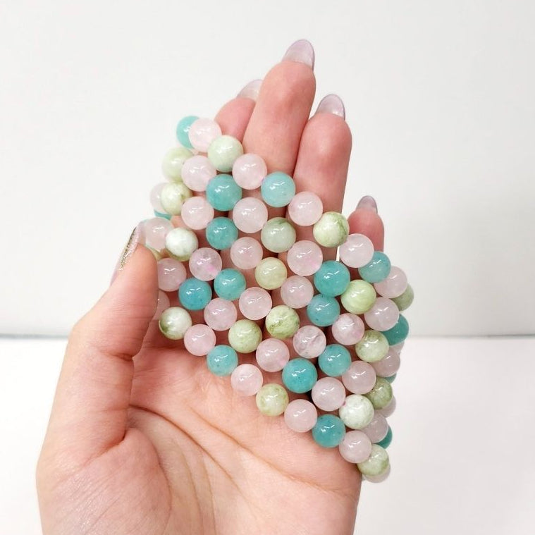 A hand holding a five Amazonite, Jade and Rose Quartz crystal bead bracelets , small round beads in pastel pink, mint green, and aqua blue, photographed against a plain white background.