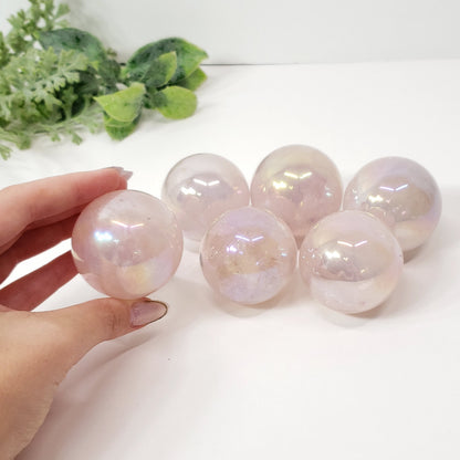 Five iridescent pale pink crystal spheres displayed on a white surface, with a hand holding one sphere in the foreground and green leaves in the upper left corner.