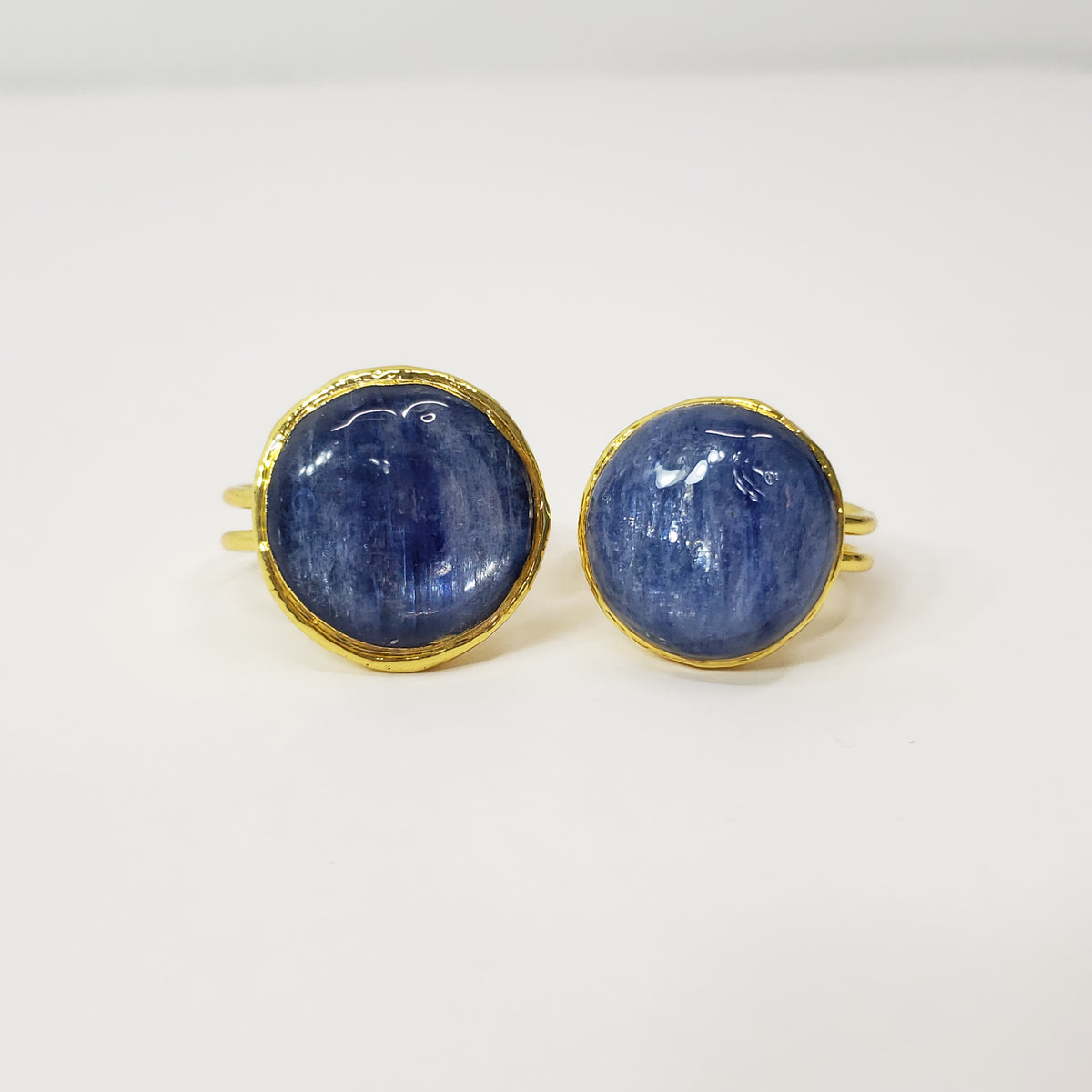 A pair of gold-toned rings with round, domed blue sapphire gemstones, displayed on a clean white surface.