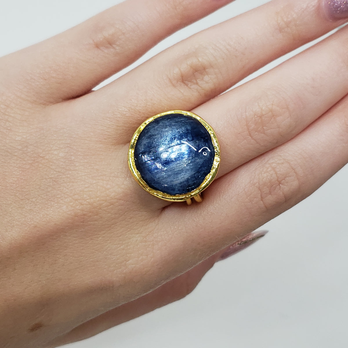 A gold ring with a large, round, deep blue kyanite cabochon gemstone featuring white star-like inclusions, worn on a person's finger.