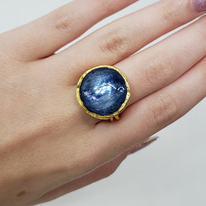 A gold ring with a large, round, deep blue kyanite cabochon gemstone featuring white star-like inclusions, worn on a person's finger.