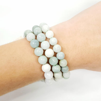 A close-up of a person's wrist wearing three beaded Burma Jade stretch bracelets made of smooth, round beads in pale sage green, white, and light gray.
