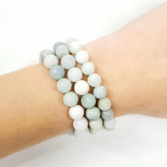 A close-up of a person's wrist wearing three beaded Burma Jade stretch bracelets made of smooth, round beads in pale sage green, white, and light gray.