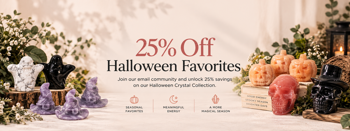 Promotional banner for the 'Halloween Favorites' collection, featuring a 25% off sale. The scene displays a variety of decorative crystal and gemstone figurines in purple, black, orange, and white, styled with small pumpkins, greenery, and candles on a white table runner against a warm beige background.