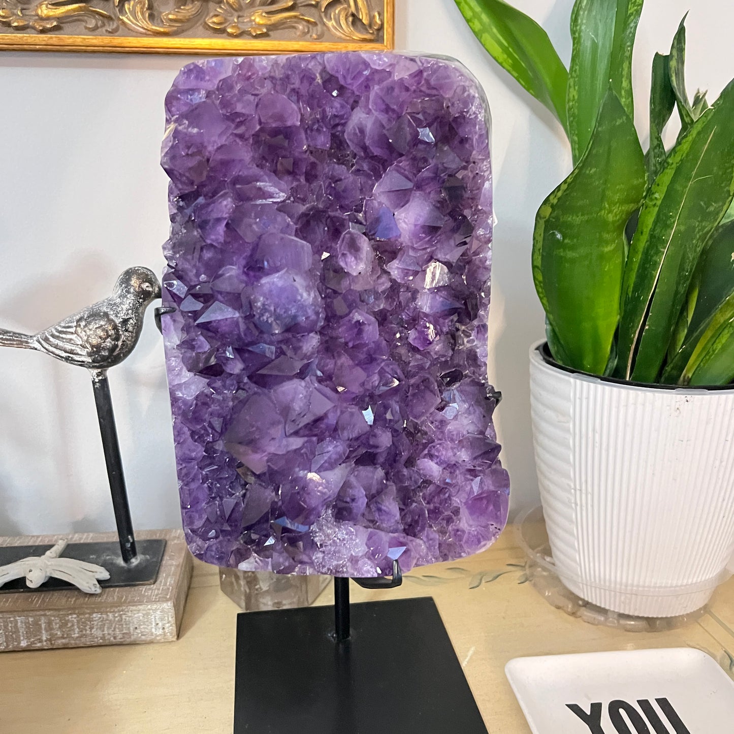 Amethyst crystal on a stand with a plant and decorative items in the background