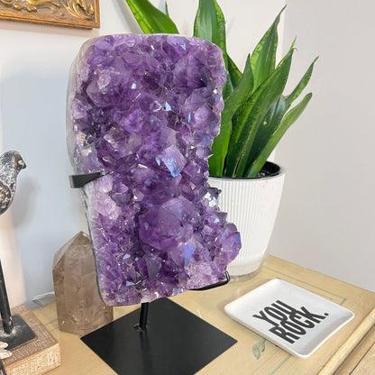 Amethyst crystal on a stand with a plant and decorative items in the background