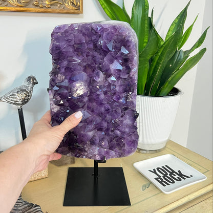 Hand held up to a large deep Purple Amethyst Geode Cluster on a stand with decorative items in the background