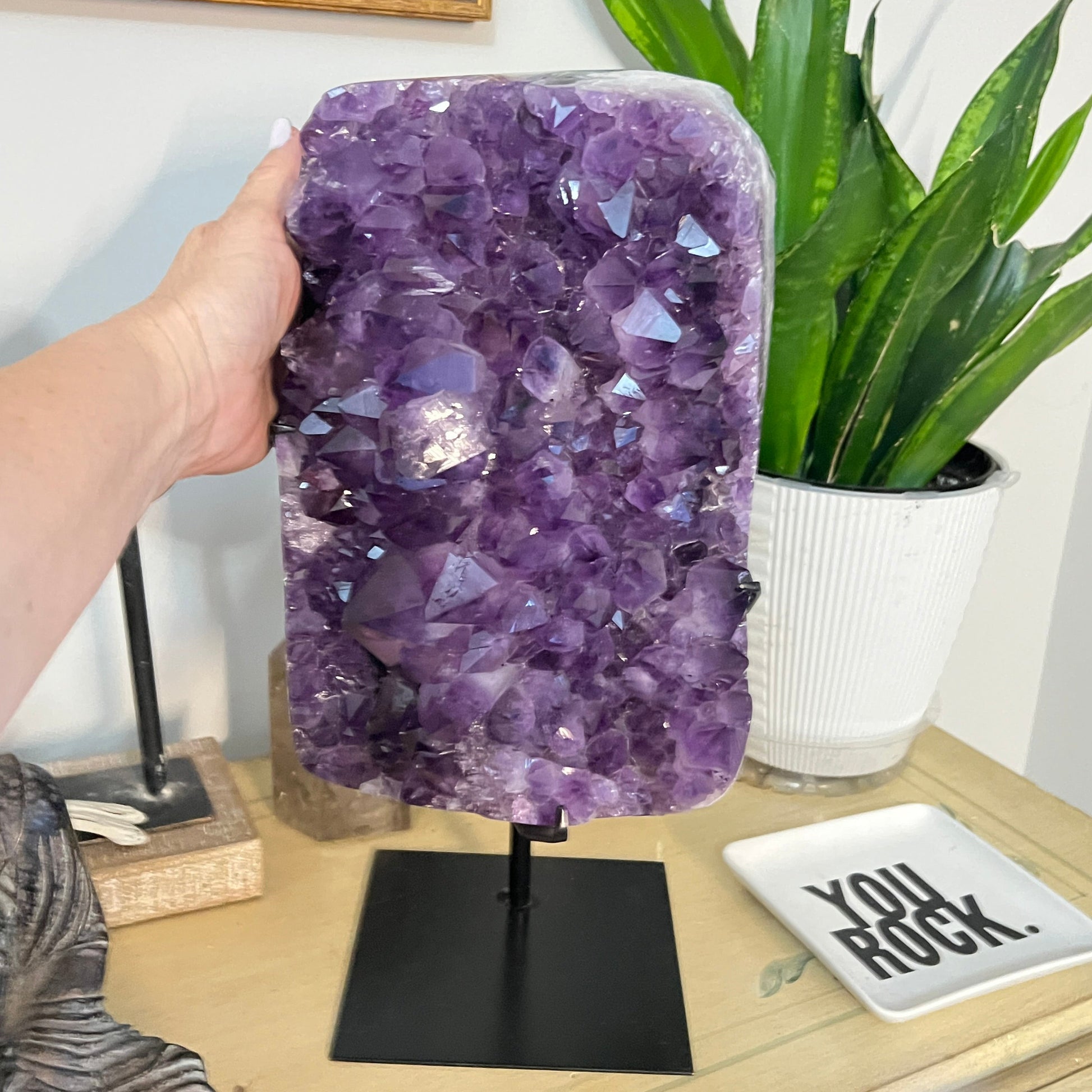 Amethyst crystal lamp on a stand with a hand holding it, plant and decorative items in the background.