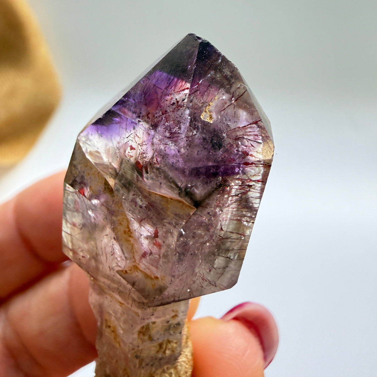 A close-up of a raw amethyst crystal point held by a hand, showing purple and lilac tones within a clear quartz structure, against a soft, out-of-focus light gray background.