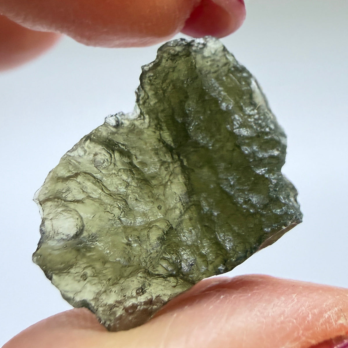 A raw, rough piece of green moldavite held between two hands with dark red painted fingernails, set against a plain light gray background.