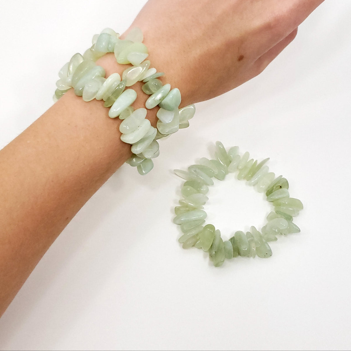 A person's hand and wrist wearing two light green jade stone bracelets, with a matching bracelet placed on a white surface below.