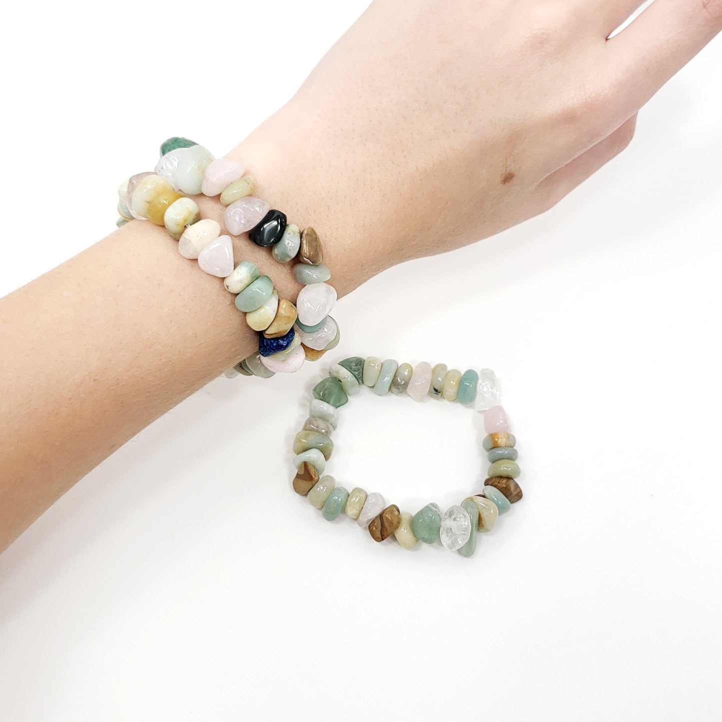 A close-up of a person's wrist wearing two beaded stretch bracelets, with a second matching bracelet placed below on a white background. The bracelets are made of small polished stones in soft pastel and earthy tones including green, pink, beige, brown, white, and a small dark accent.