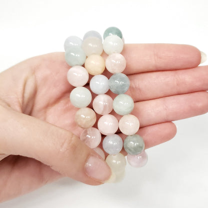 A hand holding three morganite beaded stretch bracelets made of smooth, polished round stones in pastel shades of pink, green, white, and pale blue against a clean white background.
