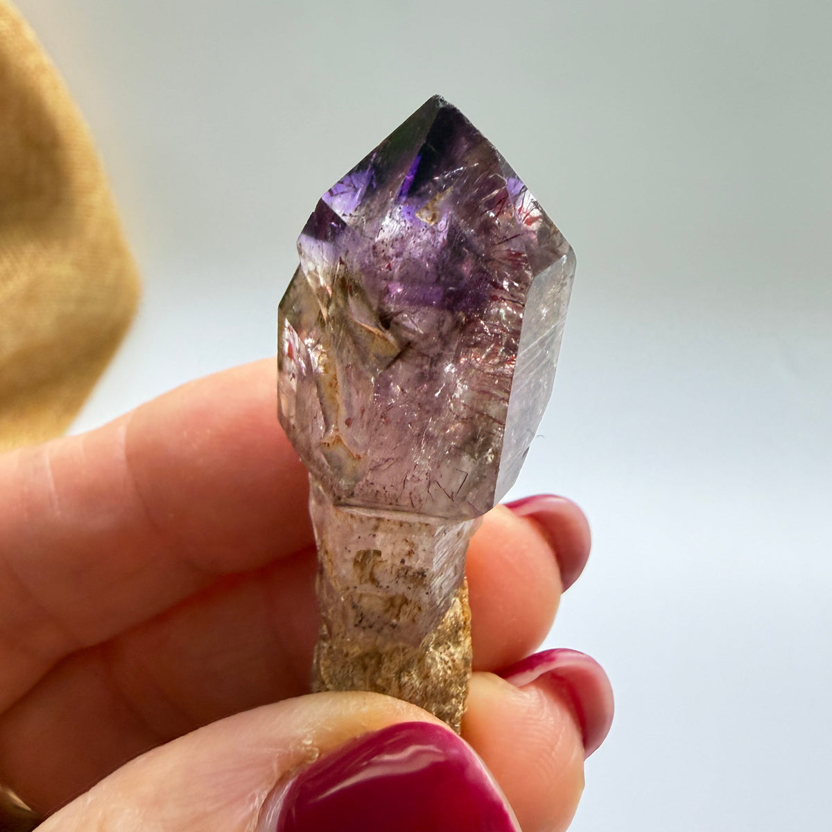 A hand with dark pink nail polish holds a raw amethyst crystal point with a rough, earthy brown base against a soft, out-of-focus light gray background.