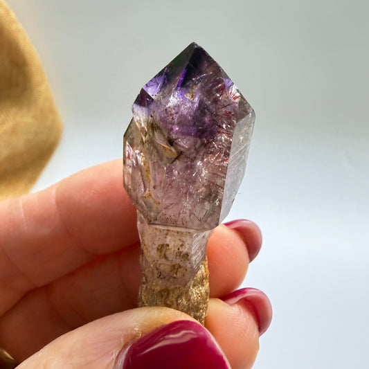 A hand with dark pink nail polish holds a raw amethyst crystal point with a rough, earthy brown base against a soft, out-of-focus light gray background.