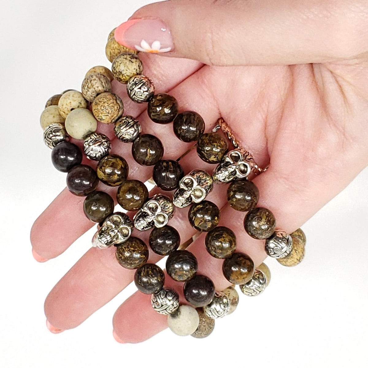 A close-up of a hand holding a picture jasper and broznite beaded bracelet. The bracelet features dark brown and black tiger's eye beads, natural beige and cream jasper beads, and metallic silver skull-shaped spacers.