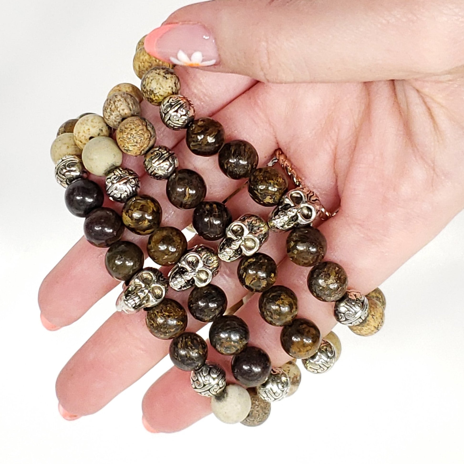 A close-up of a hand holding a picture jasper and broznite beaded bracelet. The bracelet features dark brown and black tiger's eye beads, natural beige and cream jasper beads, and metallic silver skull-shaped spacers.