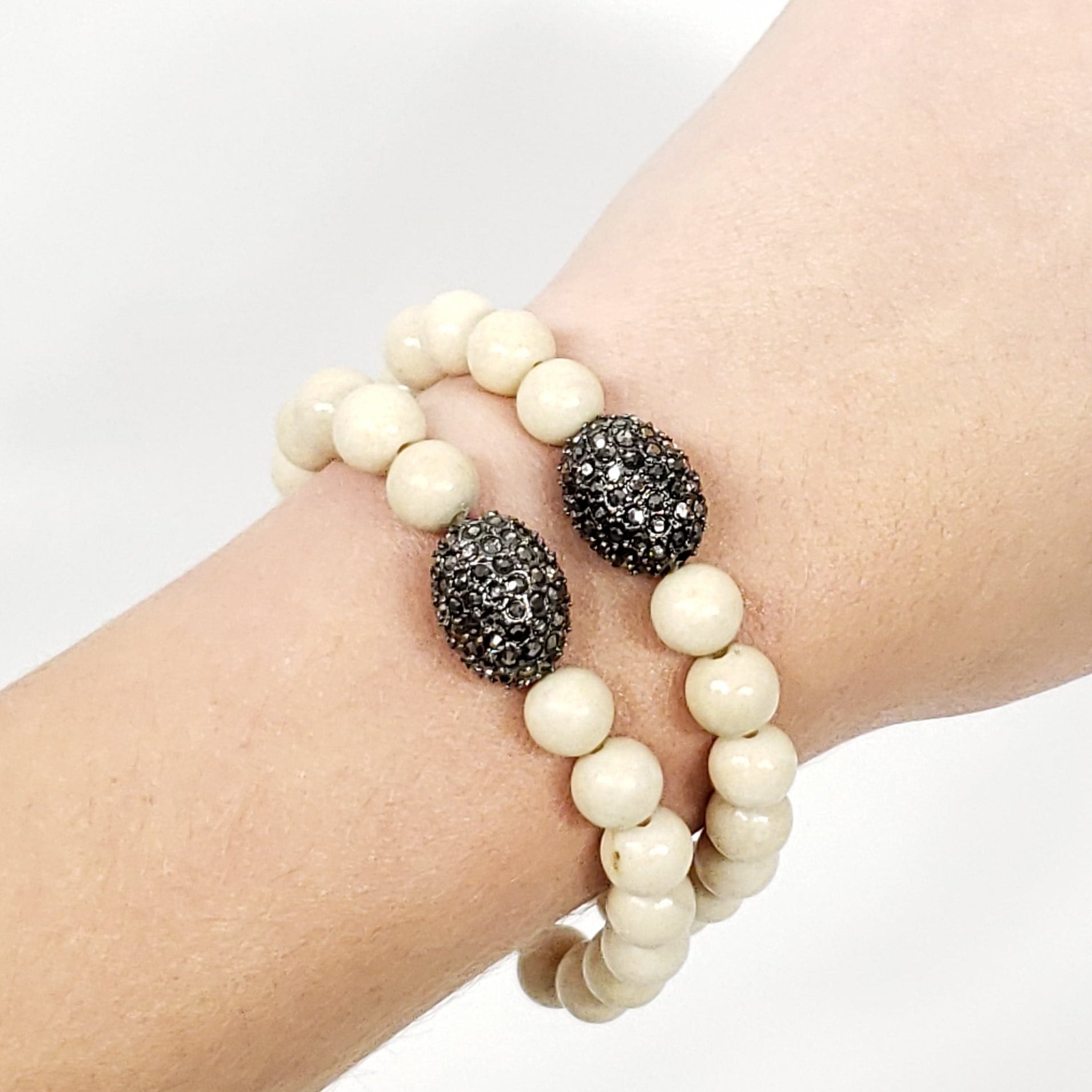 A close-up of a person's wrist wearing two riverstone jasper beaded bracelets. The bracelets consist of cream-colored round beads and two dark, textured oval beads with a metallic silver glittery finish.