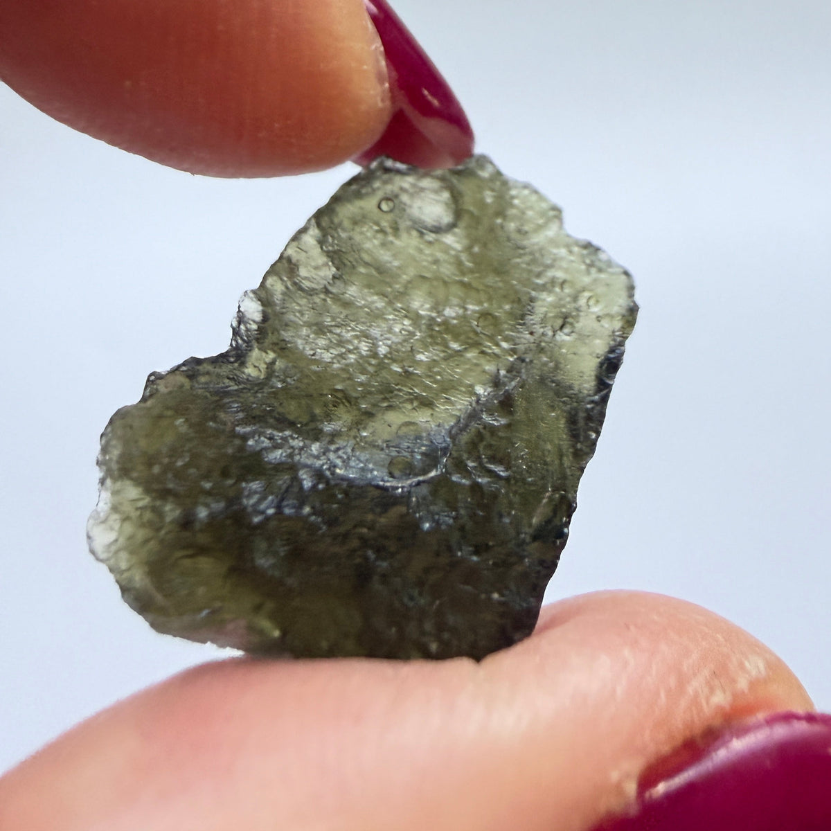 A close-up of a rough, unpolished dark green stone or mineral being held between two fingers with dark red painted nails against a plain light gray background.