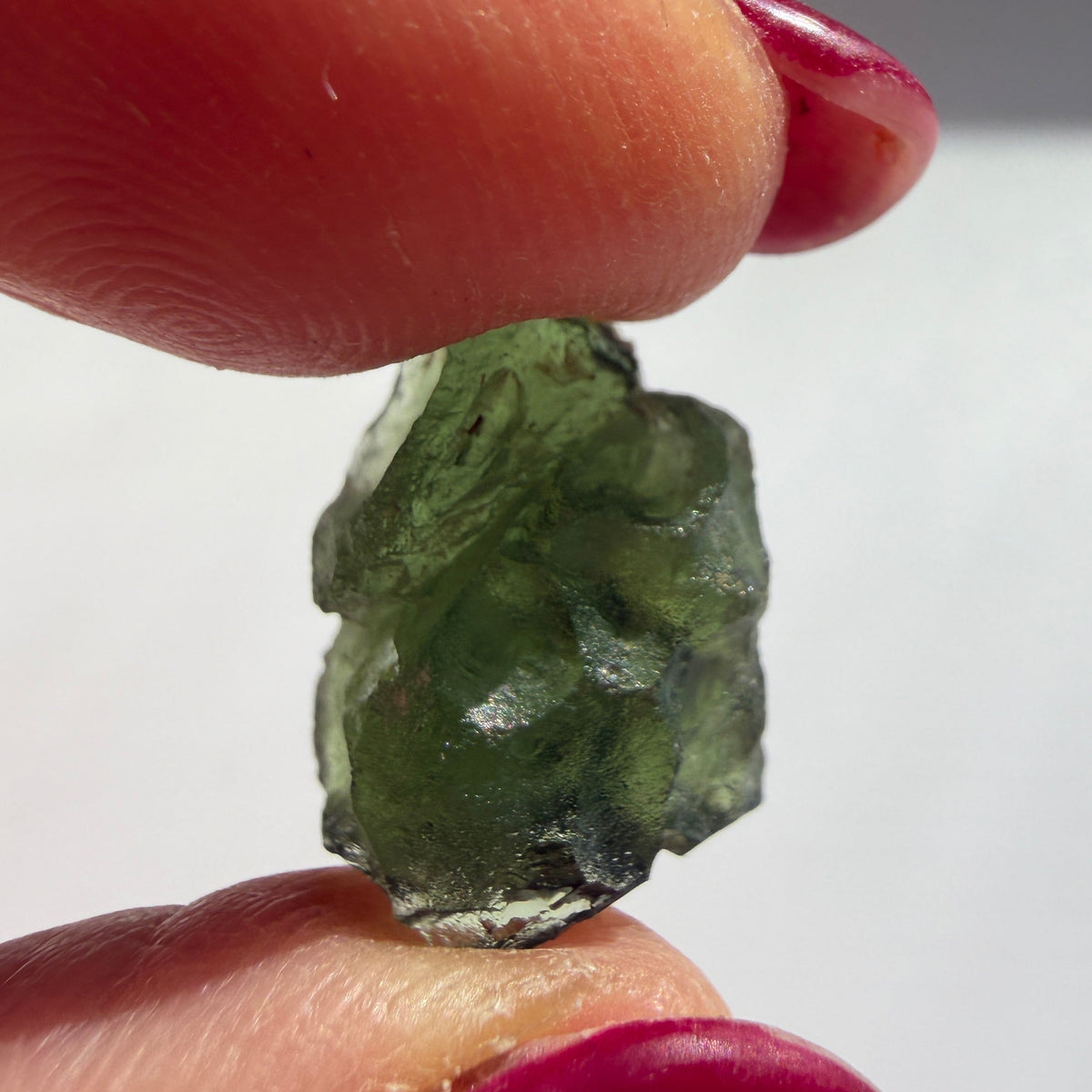 A small rough green translucent gemstone or crystal held between two fingers with dark red painted nails, against a plain light background.