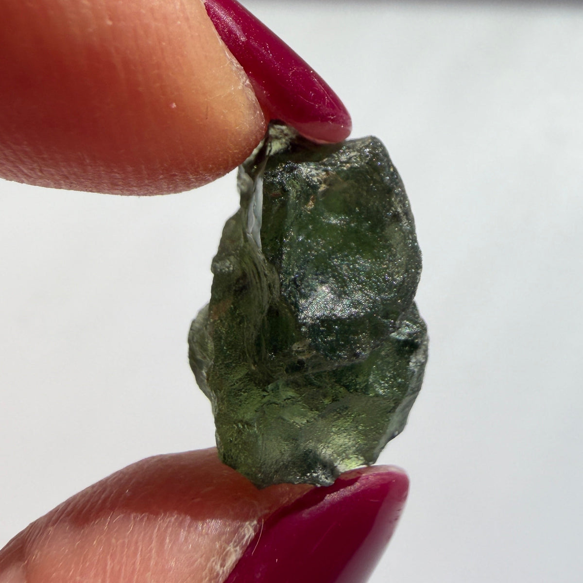 A small rough green gemstone or mineral chunk held between two fingers with dark red painted nails against a plain light background.