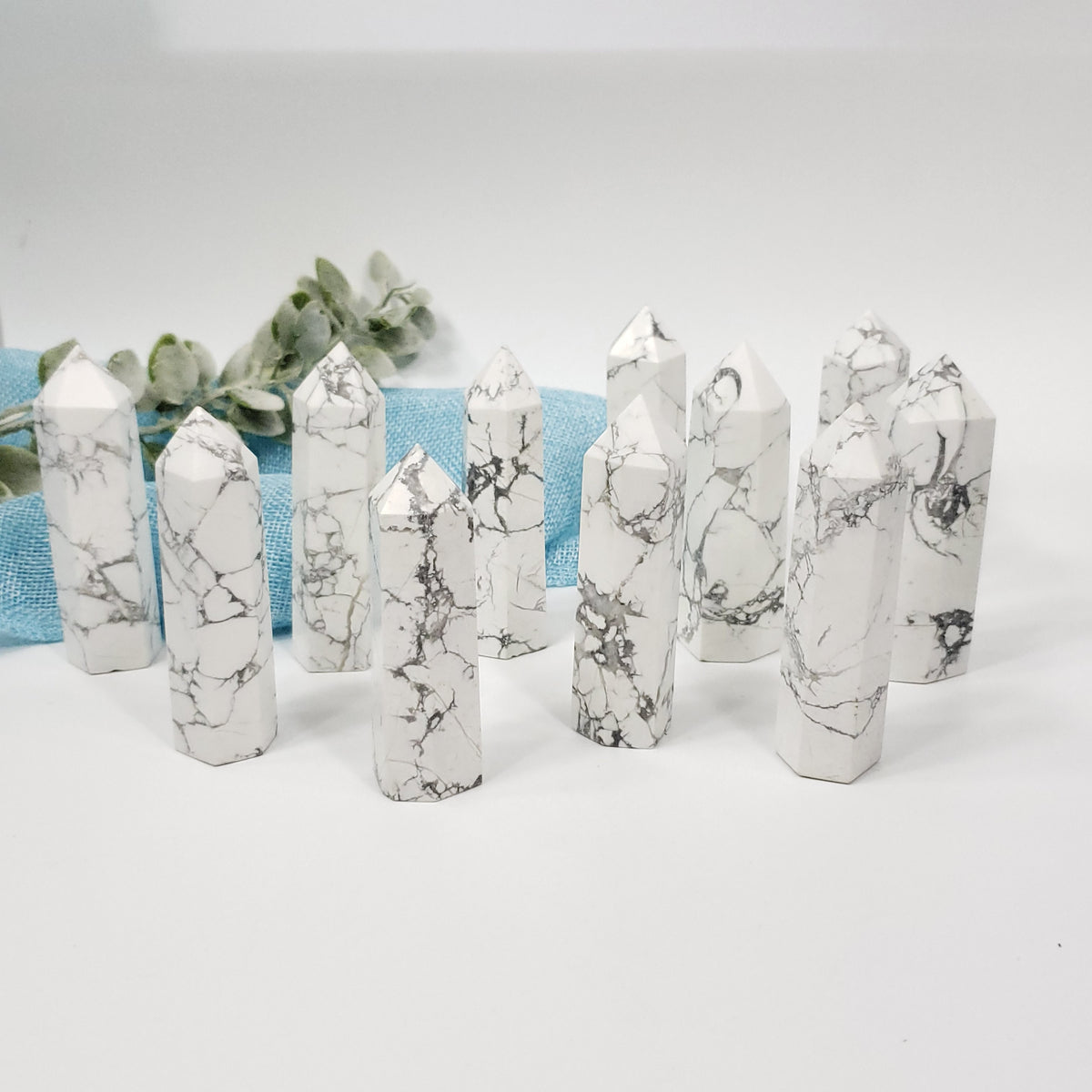 A set of white marble crystal points with gray veining, arranged on a white surface with a light blue textured fabric and green leaves in the background.