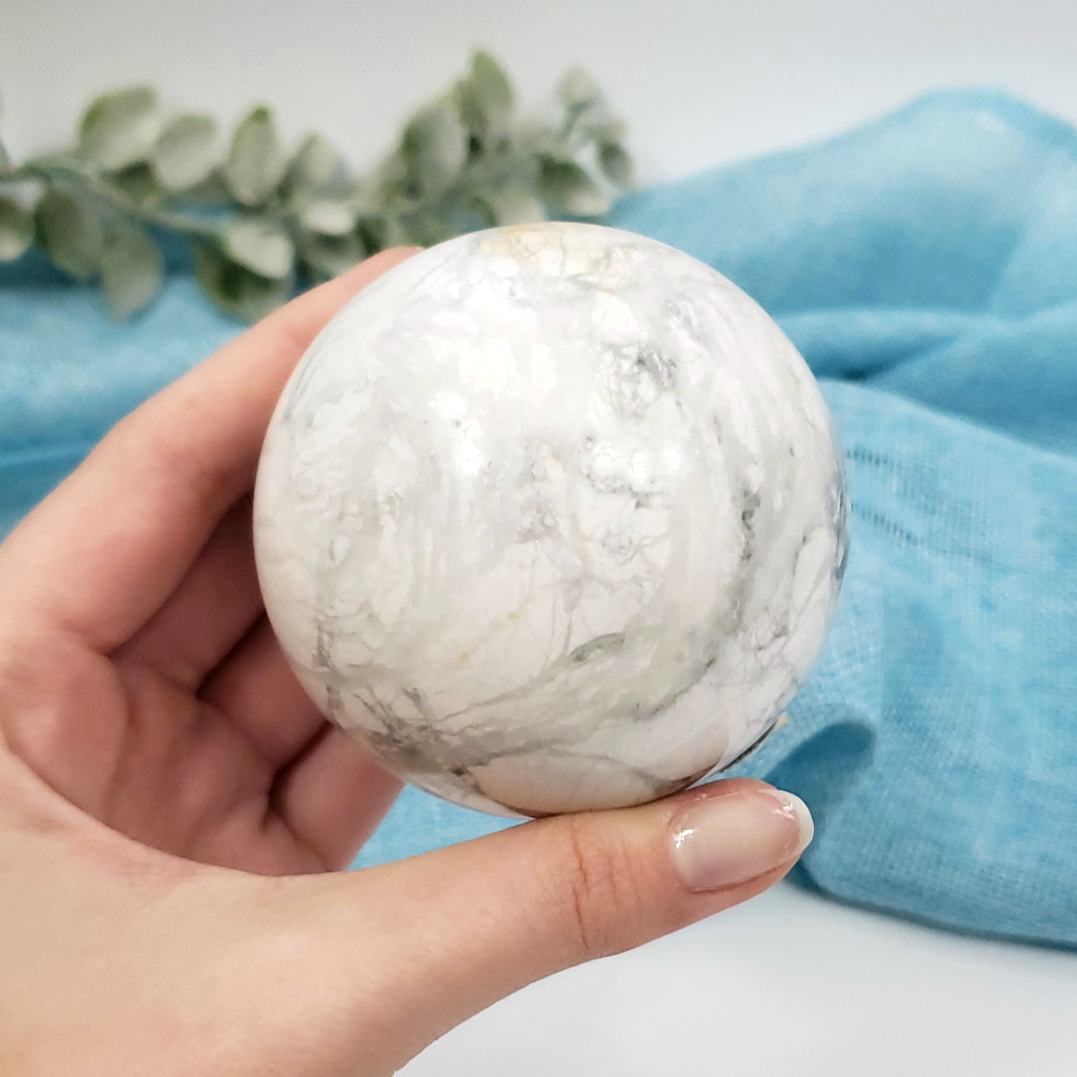 A hand holding a polished white marble sphere with gray veining, set against a soft blue fabric backdrop with a green eucalyptus branch in the background.