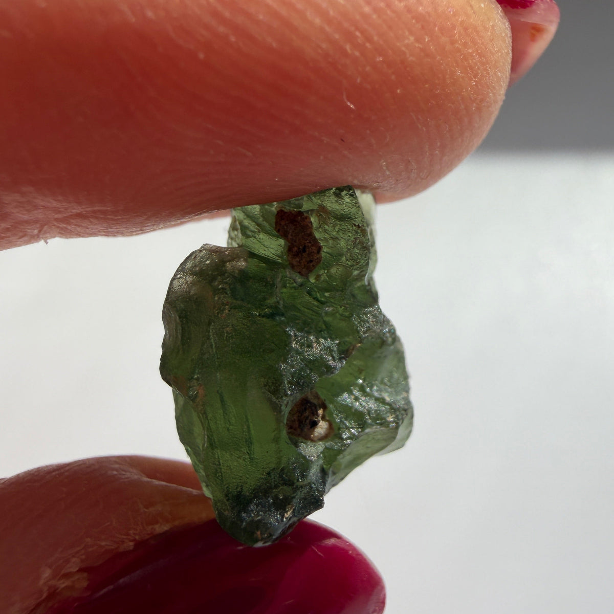A close-up of a rough, raw green gemstone held between two fingers with dark red painted fingernails against a plain light gray background.