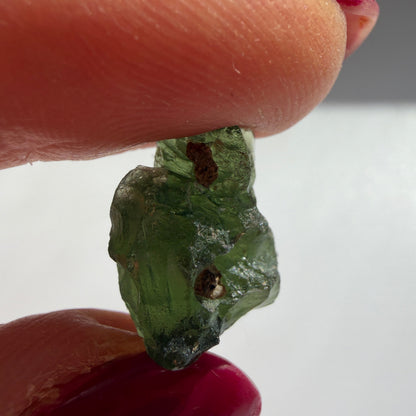 A close-up of a rough, raw green gemstone held between two fingers with dark red painted fingernails against a plain light gray background.