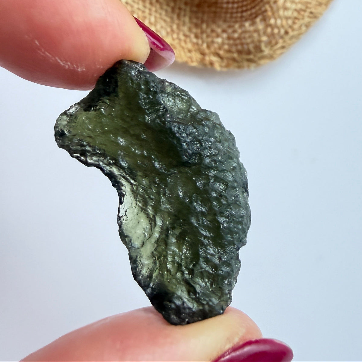 A hand with dark red painted nails holds a small, rough, dark green translucent stone or crystal against a light gray background, with a beige woven fabric pouch blurred in the upper right.