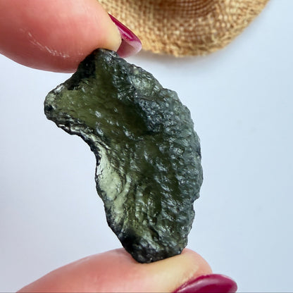 A hand with dark red painted nails holds a small, rough, dark green translucent stone or crystal against a light gray background, with a beige woven fabric pouch blurred in the upper right.