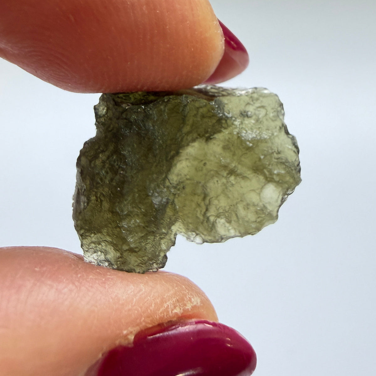 A close-up of a person's fingers with dark red painted nails holding a small, rough, translucent olive-green stone or mineral against a plain light background.