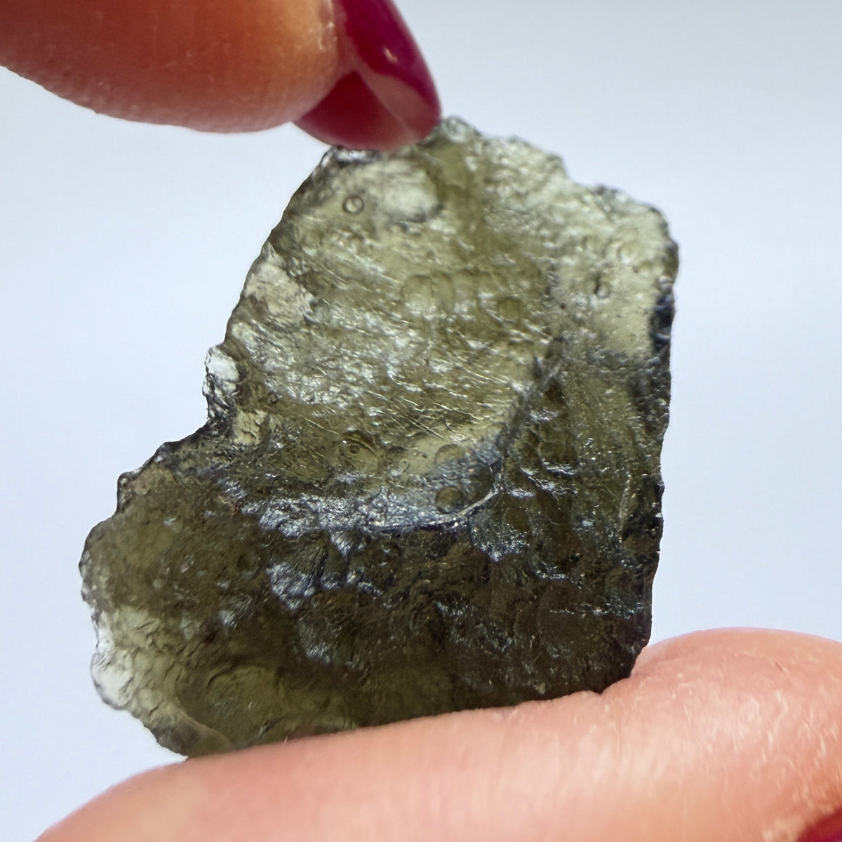 A close-up of a hand with dark red manicured nails holding a small, rough, translucent olive-green stone or crystal against a plain light background.
