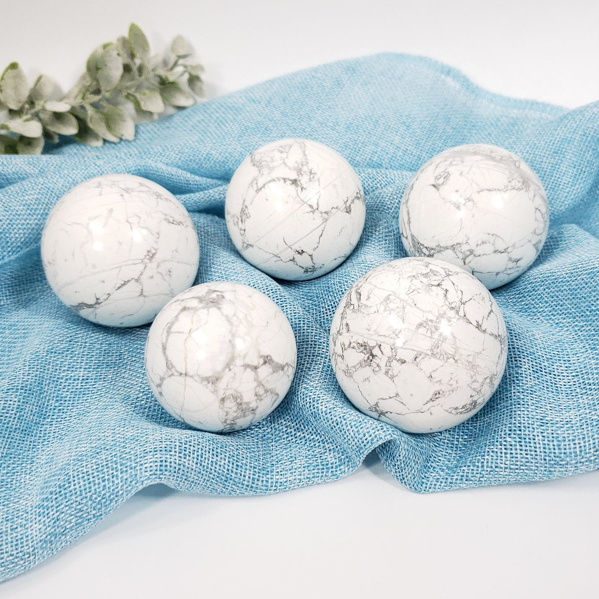 Five white marble-like d, white howlite cyrstal spheres  arranged on a light blue textured fabric surface with a small leafy sprig in the corner.