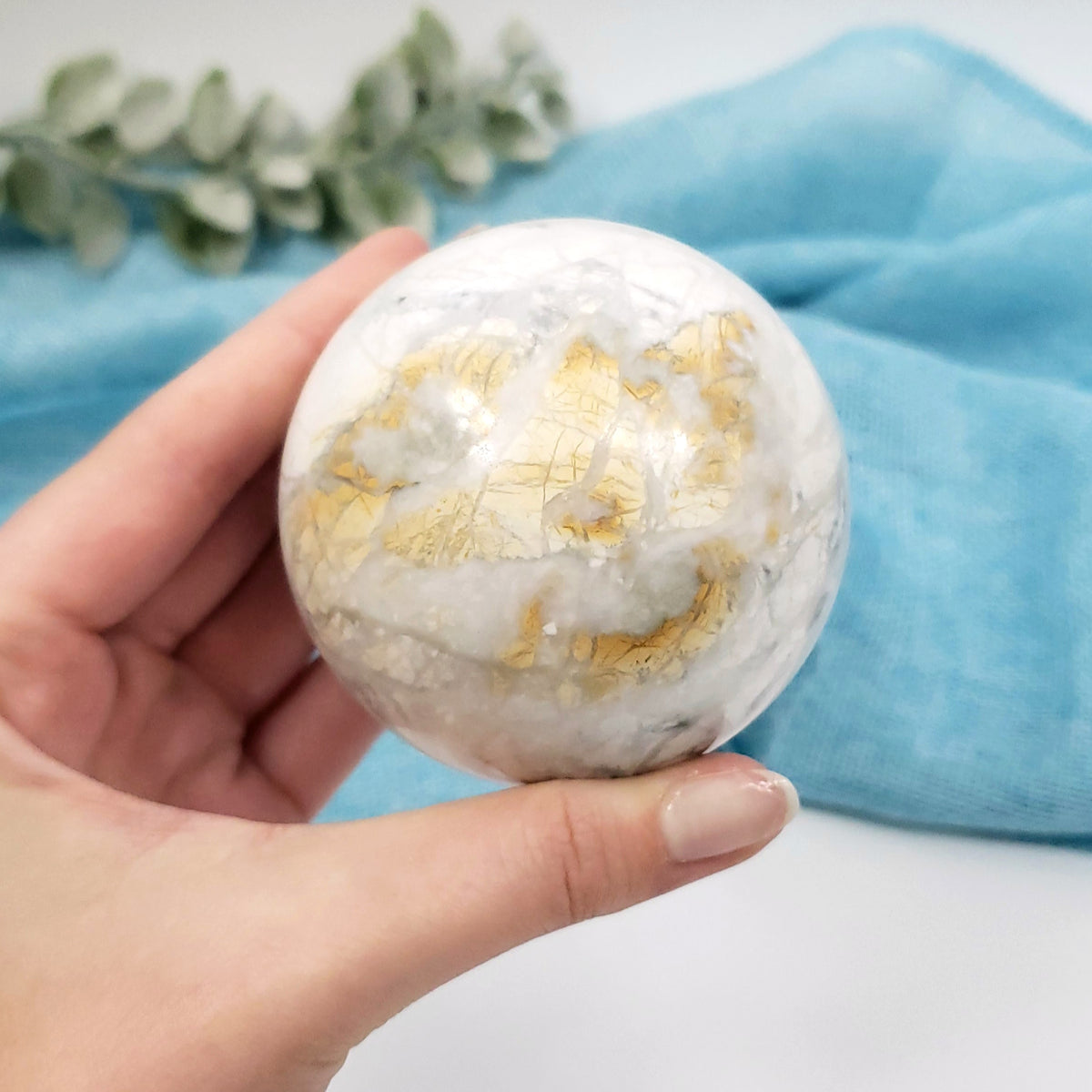 A hand holding a polished white and gold marble sphere, with a soft blue fabric and green leaves in the background.