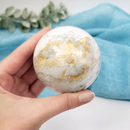 A hand holding a polished white and gold marble sphere, with a soft blue fabric and green leaves in the background.
