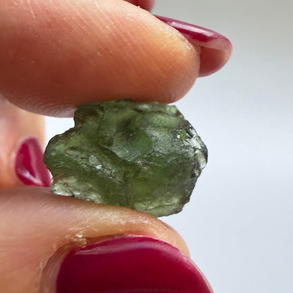 A close-up of a hand with dark red manicured nails holding a small, rough, translucent green stone or crystal against a plain light gray background.