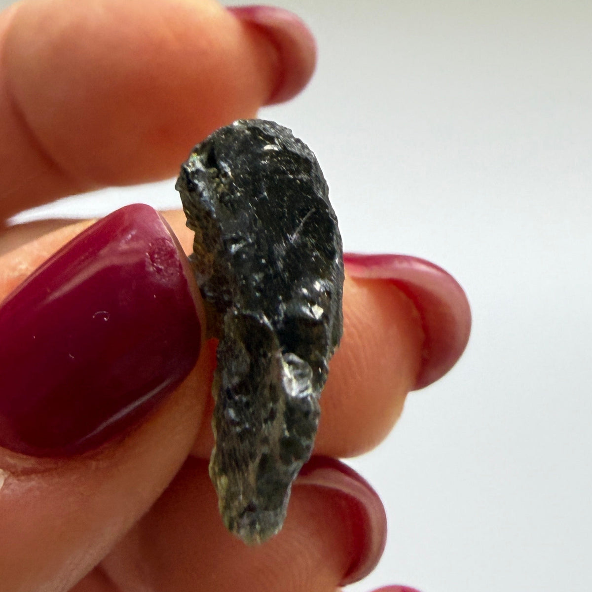 A hand with dark red painted fingernails holds a small, rough, dark green gemstone against a plain, light background.