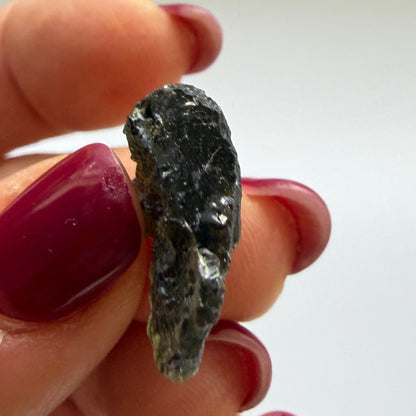 A hand with dark red painted fingernails holds a small, rough, dark green gemstone against a plain, light background.