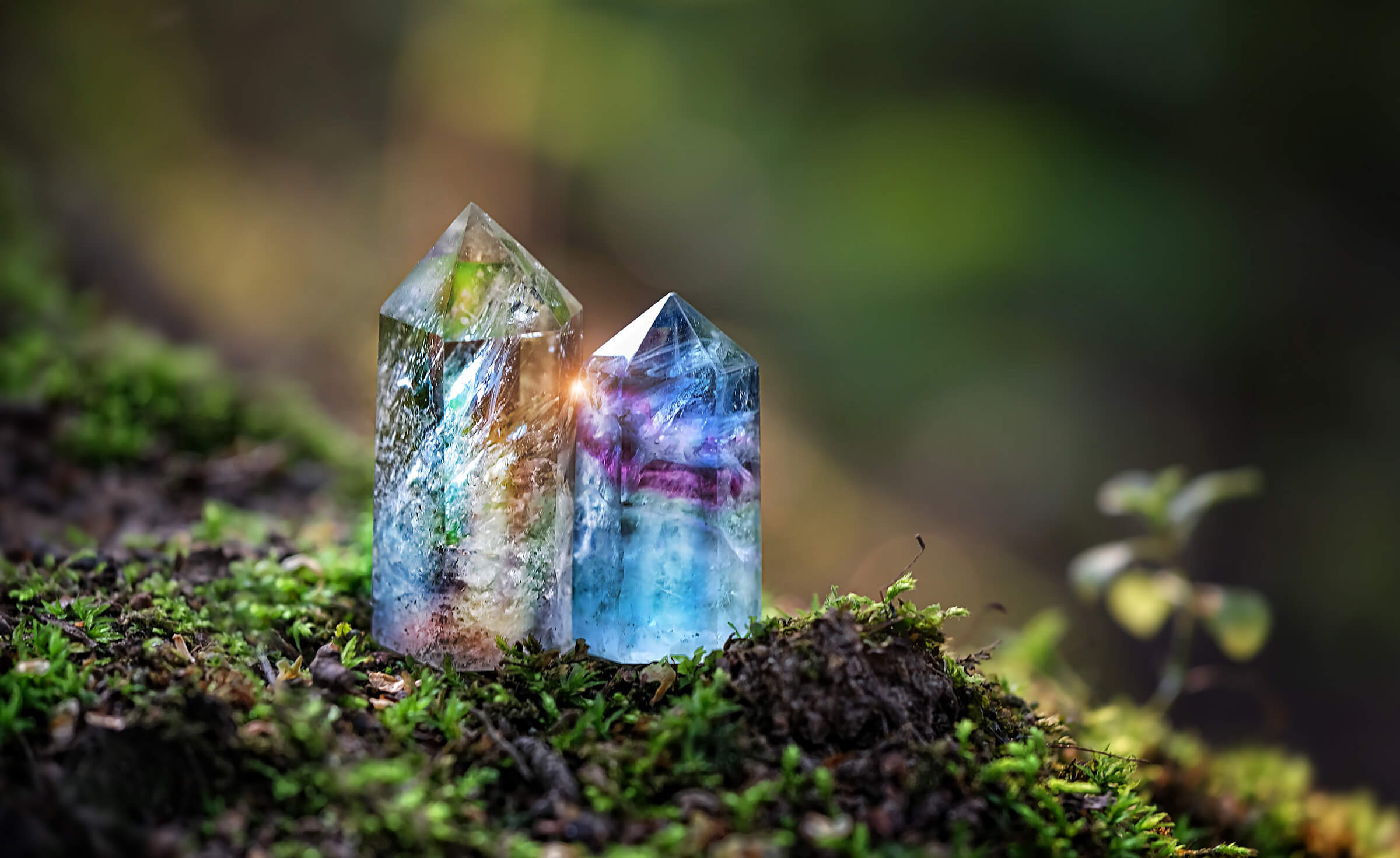 An ethically sourced clear crystal tower is alongside a purple and blue crystal tower in a natural setting
