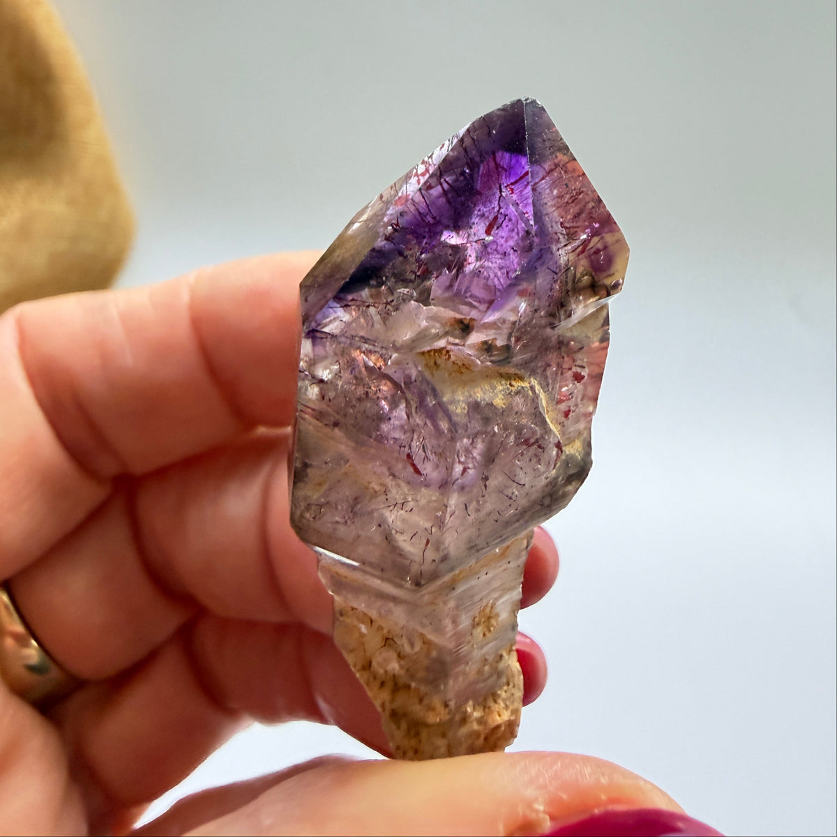 A hand holding a rough purple amethyst crystal point against a soft, light gray background.