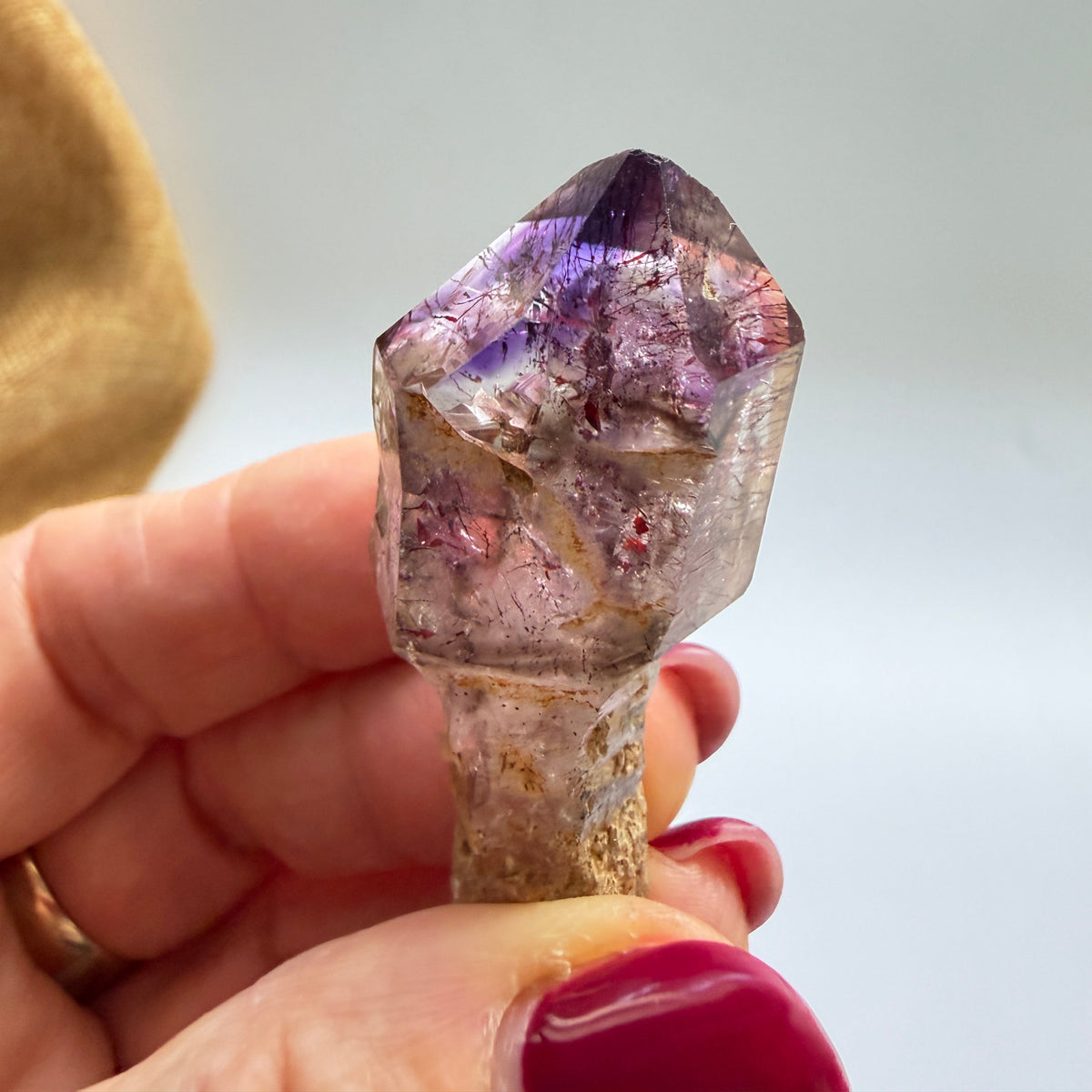 A hand with dark red painted fingernails holds a raw amethyst crystal point against a plain light background. The crystal shows natural purple and pinkish tones with some brownish matrix.