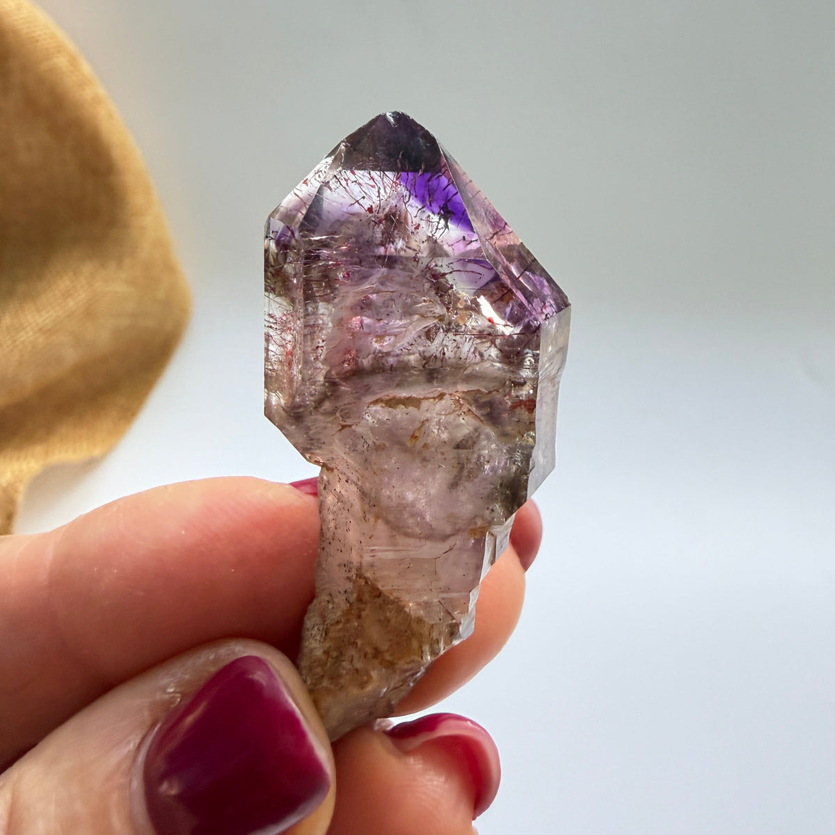A hand with dark red painted nails holds a raw amethyst crystal point against a plain light background.
