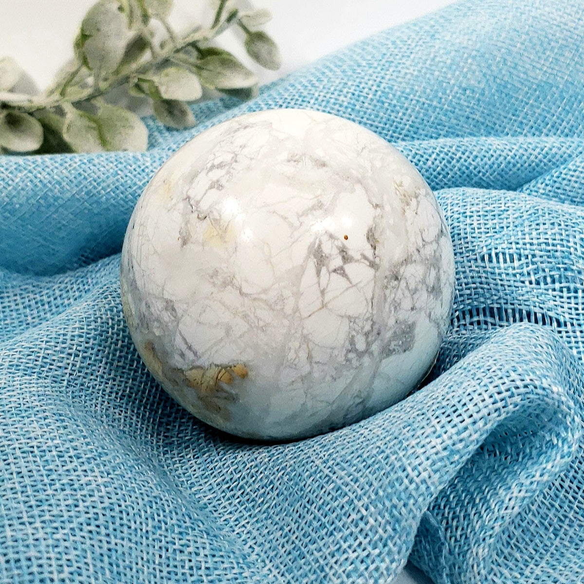 A polished white howlite, marble-like sphere with gray veining and subtle gold flecks, resting on draped light blue fabric with a small green leafy sprig in the background.
