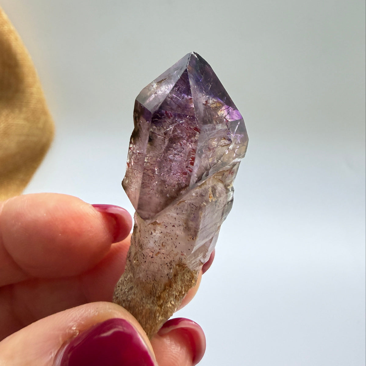 A hand with magenta nail polish holds a raw purple amethyst crystal point against a soft, out-of-focus light gray background with a tan cloth visible on the left.